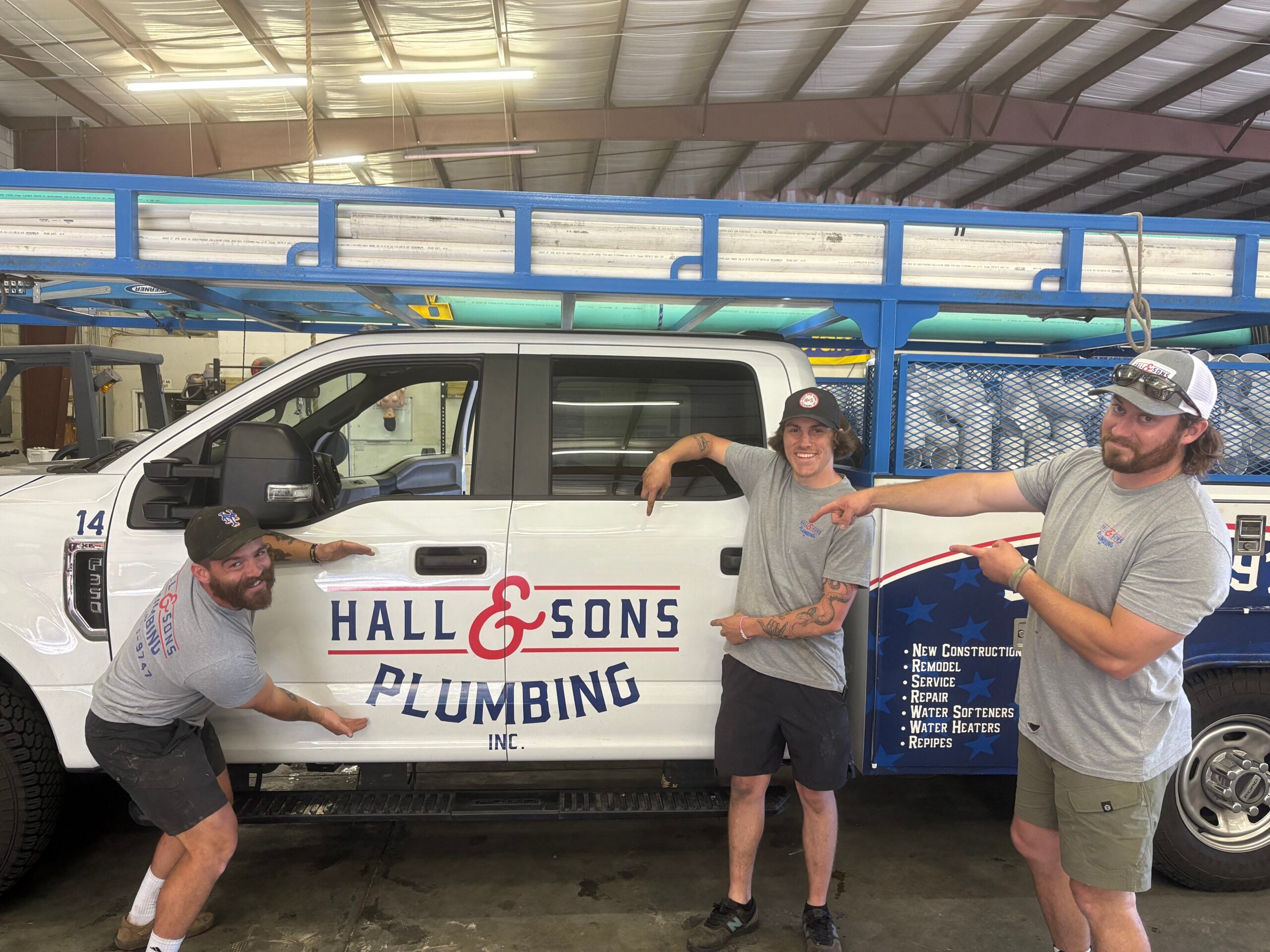 Three Hall & Sons Plumbing team members pointing at a company service truck inside a workshop/garage. The white truck features the Hall & Sons Plumbing logo and lists services including new construction, remodels, repairs, water softeners, water heaters, and repipes.