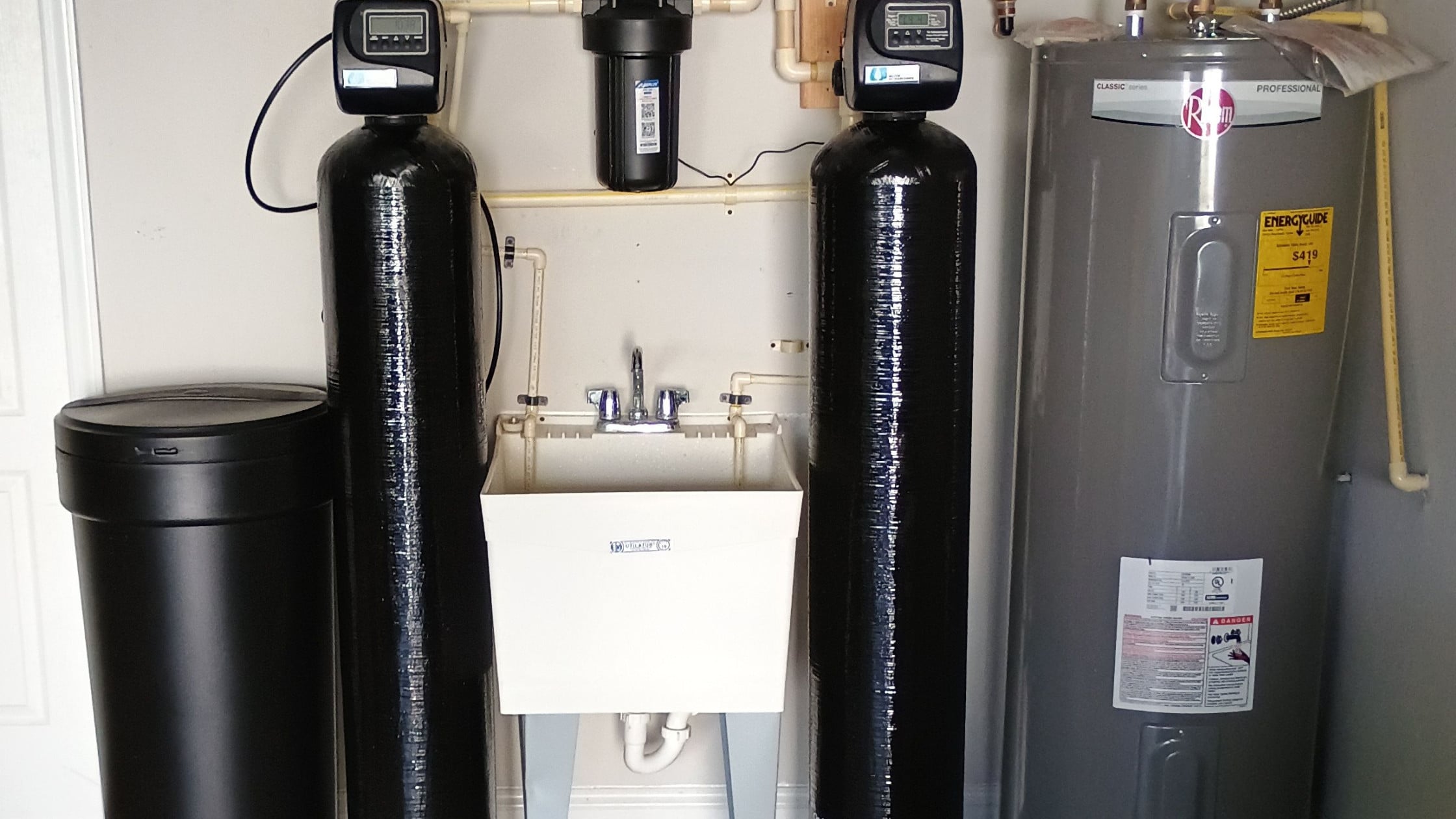 Utility room with a laundry sink flanked by two tall black water-treatment tanks, a black brine tank on the left, and a gray water heater on the right.